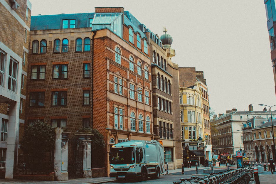 A city street scene featuring a variety of multi-storey buildings with diverse architectural styles, including a prominent red-brick structure with arched windows and decorative stonework, alongside a yellow, ornate building with intricate window frames and a rounded turret topped with a weather vane. The street is lined with parked bicycles and traffic bollards, with a grey rubbish collection vehicle from Waste Removal Belgravia positioned in the foreground, ready for waste pick-up. The vehicle is clean and modern, with a compact front cab and a larger waste storage area, suggesting a focus on efficient, private rubbish collection services in an urban environment. The environment is well-lit with natural daylight, and the scene captures a typical cityscape with adjacent buildings, street furniture, and minimal pedestrian activity, emphasizing the importance of on-site waste clearance and alternative waste handling options for residential or commercial properties in the area.