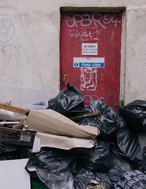 An outdoor scene showing a large pile of black rubbish bags and miscellaneous waste materials, including cardboard and plastic, stacked against a textured, off-white building wall. In the background, there is a small, rectangular red door with graffiti markings and several signs attached, including one with the text 'No Money' and another blue sign saying 'Keep clear.' The rubbish appears to be heaped directly at the base of the door, indicating an on-site waste collection or clearance activity. The environment is outdoors with natural lighting, and the waste materials look disorganized, typical of private rubbish removal services such as those provided by Waste Removal Belgravia, supporting the context of alternative waste handling arrangements in urban settings.