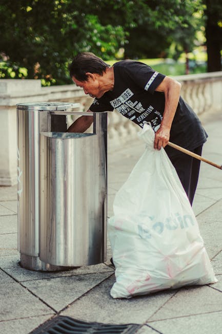 A middle-aged woman wearing a short-sleeved black T-shirt with printed text and dark trousers is seen disposing of waste into a metallic recycling bin located on a paved sidewalk. She is bent over slightly, holding a large white plastic bag filled with rubbish in her left hand, while her right hand is reaching towards the top of the bin. The bin is cylindrical with a brushed metal finish, positioned next to a concrete railing that runs along a park or garden area. The background features lush green trees and foliage, with sunlight filtering through the leaves, creating a natural outdoor setting. The scene is clean and orderly, capturing a moment of private waste disposal in a public space, which aligns with concepts of independent collection or on-site rubbish management. Waste Removal Belgravia's service focus on rubbish removal is subtly supported by this visual depiction of waste being prepared for collection, emphasizing environmental responsibility and professional waste handling.