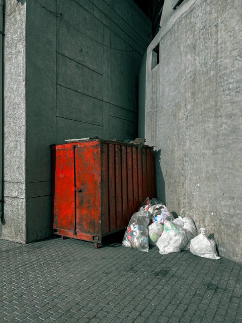 A red metal skip with a weathered and rusty surface is positioned against the corner of a dark green wall and a light grey concrete wall on a paved sidewalk. The skip is partially filled with trash, and several clear plastic bags of waste, containing mixed rubbish such as paper, plastic bottles, and other household waste, are piled on the ground next to it. The scene appears to be an urban outdoor area set on a grey paving surface, with no visible signage or other street furniture. Natural daylight illuminates the scene, highlighting the textures of the rusted metal, the smooth concrete, and the plastic waste. This image represents a typical privately managed waste collection point, aligned with the concept of independent rubbish removal services that complement municipal waste systems, with Waste Removal Belgravia providing such solutions in the local area.