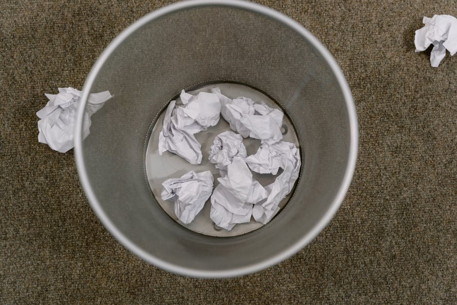 An overhead view of a cylindrical grey metal bin positioned on a brown textured carpeted floor, partially filled with crumpled white paper waste and standing on a flat surface. Several loose crumpled paper balls are scattered on the carpet around the bin, indicating a typical internal office or residential waste collection setting. The bin appears to be lined with a plastic bag visible around the edges, and the crumpled paper inside suggests typical paper waste from desks or informal disposal. The environment around the bin is free of other objects, with natural ambient lighting creating soft shadows, and the focus is on the waste contained within and around the bin. This scene illustrates a common example of a stationery waste collection point, relevant to private rubbish removal services such as those provided by Waste Removal Belgravia, for efficient on-site clearance or alternative waste handling in a professional setting.