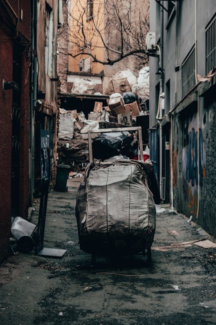 The image depicts a narrow urban alleyway filled with a large accumulation of mixed waste and debris, including cardboard boxes, plastic bags, and assorted refuse piled high against a back wall. In the foreground, a large, weathered grey wheeled rubbish bag with visible creases and dirt stains is positioned centrally on the uneven, grimy asphalt surface. Surrounding the scene are tall, closely spaced buildings with graffiti on the lower sections of the walls, metal railings, and exposed pipes, indicating a neglected and cluttered environment. The background features a pile of garbage stacked beneath leafless tree branches and an overcast sky, casting subdued, diffuse lighting over the scene. On the right, a grey metal fence and a small utility box are attached to the building façade. The setting suggests a location targeted for independent waste collection, managed by waste removal professionals such as Waste Removal Belgravia, emphasizing a private approach to rubbish disposal in a densely urban context familiar to service areas in Belgravia.