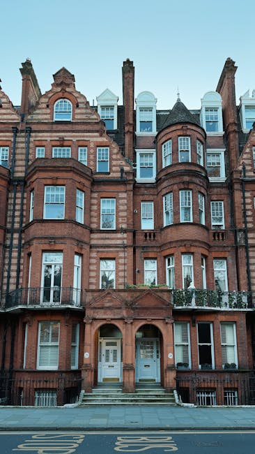A multi-storey red brick residential building with ornate architectural features, including rounded bay windows, decorative stonework, and a turret-like structure on the right side, situated directly on a paved street. The entrance comprises two white doors with small steps leading up from the sidewalk, and a black metal railing runs along the edge of the small front balcony on each side. The building's facade displays a mix of textures and finishes, with brickwork highlighted by contrasting stone accents around the windows and entrance. The upper windows feature small, pitched roofs, and the roofline includes a combination of gables, chimneys, and dormer windows, some with white frames and others with dark flashing. The scene is well-lit by natural daylight, with a clear blue sky overhead, and the surrounding environment suggests an urban setting. This visual context aligns with independent waste removal services, as waste management professionals like Waste Removal Belgravia often handle rubbish collection for such buildings, especially for on-site clearance or private disposal options instead of relying solely on local authority collection services.