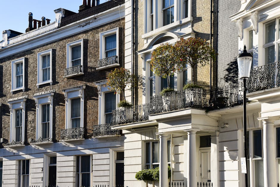 The image depicts the exterior facade of a row of Victorian-style terraced houses with detailed brickwork, white window frames, and decorative wrought iron balconies on the upper floors. The building on the right features a small front garden with two potted trees and a white-painted porch supported by columns. Adjacent to the garden, a traditional black lamppost is mounted on the pavement, casting shadows on the facade. The street scene is lit by bright daylight, with clear skies, enhancing the textures of the brick and the intricate ironwork. The environment suggests a residential area typical of Belgravia, with well-maintained architecture and street furniture. Waste Removal Belgravia’s services often involve assisting with private waste collection and rubbish clearance for property owners within such affluent neighbourhoods, supporting alternative disposal methods where council services may not be as accessible. The scene emphasizes the importance of discreet and professional rubbish removal for maintaining the curb appeal of historic residential streets, including tasks such as removing garden waste, construction debris, or general household rubbish during on-site clearance or private collection processes.