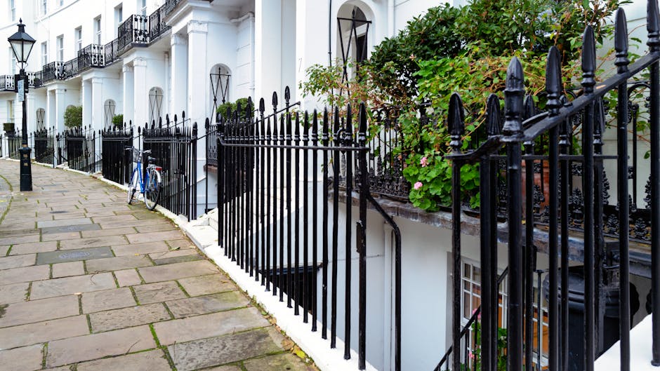 A row of white terraced townhouses with black wrought iron fencing along a concrete and stone-paved sidewalk in a residential area. The fencing includes vertical bars with pointed finials at the top and is positioned in front of small front gardens that feature greenery and flowering plants. One of the front gardens has a pink-flowering shrub visible behind the fence, while another has neatly trimmed bushes. A blue bicycle is parked and leaning against the fence near the pavement. The buildings have large windows, some with black decorative ironwork, and potted plants on the window sills. The scene is well-lit with natural daylight, highlighting the textures of the wrought iron, the smooth white facade of the buildings, and the cobblestone pavement. This setting demonstrates a typical Belgravia residential street, highlighting options for private waste management and on-site clearance, which Waste Removal Belgravia provides to implement alternative waste handling methods for flats and townhouses in the area.